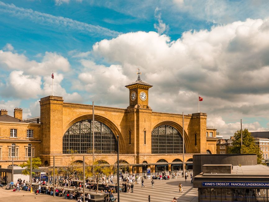 A large, historic railway station with a prominent central clock tower topped by a weather vane, built from golden-brown bricks. The station features arched windows and an arched entrance, with flags 