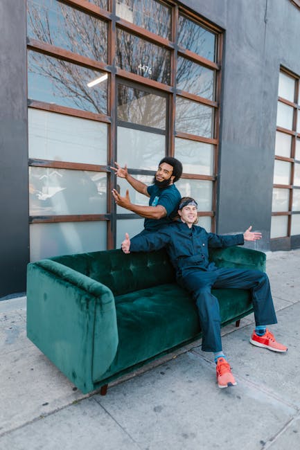 Two young men are sitting on a luxurious, emerald green velvet sofa placed outside a modern building with large wooden-framed windows. One man, wearing a navy blue jacket and matching pants with brigh