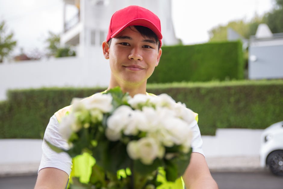 A young delivery person wearing a red cap and a high-visibility vest is holding a bouquet of white flowers, which includes roses and other blooms, wrapped in clear plastic. He is standing outdoors on 